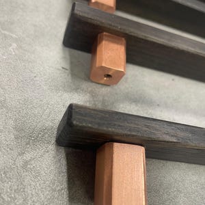 May include: Close-up of three dark wood bars with copper-colored, hexagonal knobs. The wood has a dark, rich grain. The copper knobs are a warm, metallic color. The objects are likely furniture hardware.