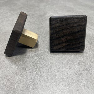 May include: Two square wooden cabinet knobs with a dark finish and brass hardware. One knob is upright, and the other is angled, showing the brass base. The knobs are likely for furniture or cabinetry.