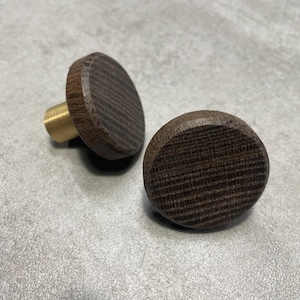以下が含まれることがあります： 木製のキャビネットノブ2つ。濃い茶色のテクスチャ加工された表面と真鍮のステムがあります。ノブは丸く、シンプルでモダンなデザインです。キャビネットや引き出しに自然なスタイルを添えるのに最適です。
