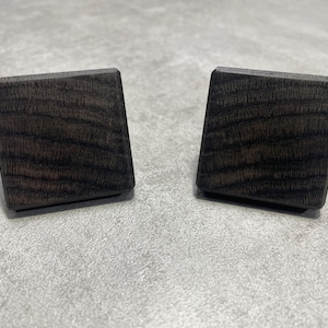 Puede incluir: Dos tiradores cuadrados de madera marrón oscuro con veta visible. Los tiradores están ligeramente angulados y se asientan sobre una superficie gris claro. Estos tiradores decorativos son adecuados para muebles.