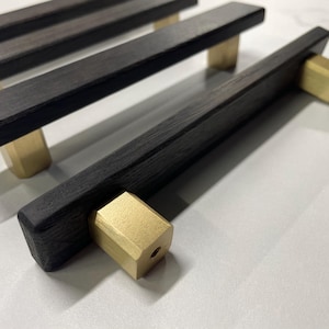 Könnte beinhalten: Nahaufnahme mehrerer dunkler Holzschrankgriffe mit quadratischen Endstücken in Messingfarbe. Die Griffe sind rechteckig und haben eine glatte, dunkle Oberfläche. Die Messingteile sind warmgoldfarben.
