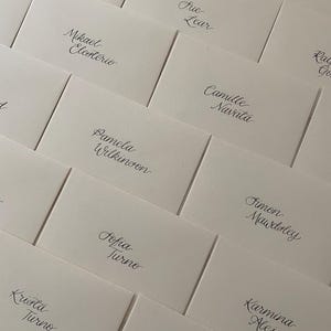 May include: An assortment of off-white name cards, arranged diagonally. Each card displays a name in stylish, black calligraphy. Names include Christina Harrio, Mikael Elesterio, Pamela Wilkinson, Sofia Turno, and others.