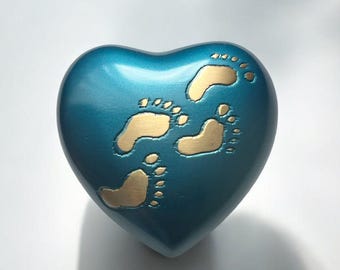 Puede incluir: Urna conmemorativa en forma de corazón de color turquesa con huellas de bebé doradas. El acabado brillante refleja la luz, resaltando las huellas grabadas. Un conmovedor recuerdo.