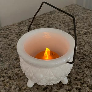 May include: A white milk glass cauldron with a textured diamond pattern, three small feet, and a thin dark metal wire handle. Inside the vessel sits a small, battery-operated orange LED tea light that emits a warm, glowing flame-shaped light.