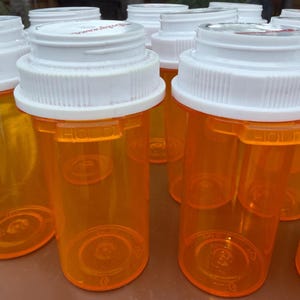 May include: A collection of empty, translucent orange plastic prescription pill bottles with white screw-top lids. Each cylindrical container features a molded plastic rim with the word HOLD embossed on the side, designed for medication storage.