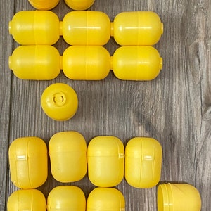 May include: A collection of bright yellow plastic capsule toys, some assembled and some disassembled, are arranged on a wooden surface. The capsules are cylindrical with rounded ends, and some have a small opening. The arrangement suggests a toy or collectible theme.