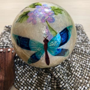 May include: A decorative sphere with a dragonfly design. The sphere features a dragonfly with blue wings and a brown body, along with purple and green floral accents. The sphere is set on a textured, beaded surface.