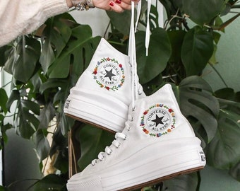 May include: White high-top Converse sneakers with a platform sole. The sneakers have a floral embroidery design around the Converse logo and the words "Chuck Taylor All Star".