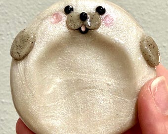 May include: A circular, off-white, shimmering ceramic dish shaped like a seal. The seal has a speckled gray snout, two black eyes, a small black nose, and pink cheeks. Two small, speckled gray fins extend from the sides of the round dish.