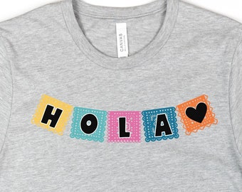 Peut inclure: Un t-shirt gris avec un motif de bannière "Hola" coloré avec un cœur. La bannière est faite de découpes de papier avec un design mexicain traditionnel. Le t-shirt est présenté sur un fond blanc avec un jean, une tasse à café et des sandales.