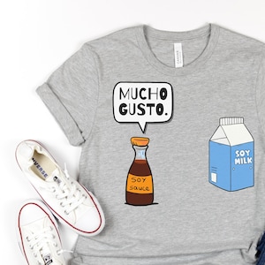 Peut inclure: Un t-shirt gris avec une illustration de dessin animé d'une bouteille de sauce soja et d'un carton de lait de soja. La bouteille de sauce soja porte l'inscription "SOY sauce" et la bulle de dialogue dit "MUCHO GUSTO." Le t-shirt est associé à un jean bleu et des baskets blanches.