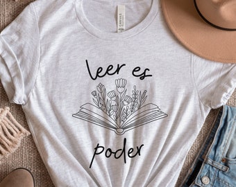 Può includere: Una maglietta grigia con il testo "leer es poder" in lettere nere, con un disegno stilizzato di un libro aperto con fiori che ne escono.