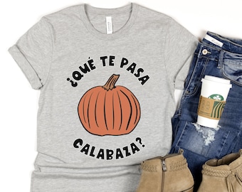 Puede incluir: Una camiseta gris con un gráfico de calabaza naranja y negro y el texto "¿Qué te pasa calabaza?" en español.