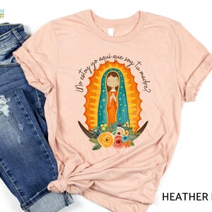 Puede incluir: Una camiseta de color melocot&oacute;n con un gr&aacute;fico de la Virgen Mar&iacute;a en un marco floral colorido. El texto "&iexcl;No estoy yo aqu&iacute; que soy tu madre?" est&aacute; impreso en la camiseta.