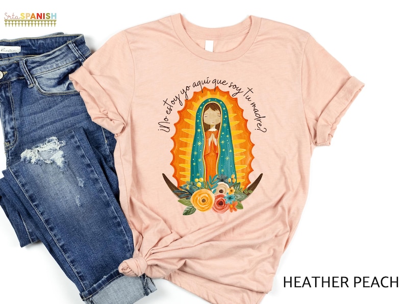 Puede incluir: Una camiseta de color melocot&oacute;n con un gr&aacute;fico de la Virgen Mar&iacute;a en un marco floral colorido. El texto "&iexcl;No estoy yo aqu&iacute; que soy tu madre?" est&aacute; impreso en la camiseta.