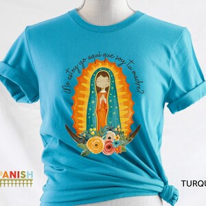 Puede incluir: Una camiseta azul turquesa con un gr&aacute;fico de la Virgen Mar&iacute;a en un halo dorado y azul. El texto "Yo estoy yo aqui que soy tu madre?" est&aacute; impreso en la camiseta.
