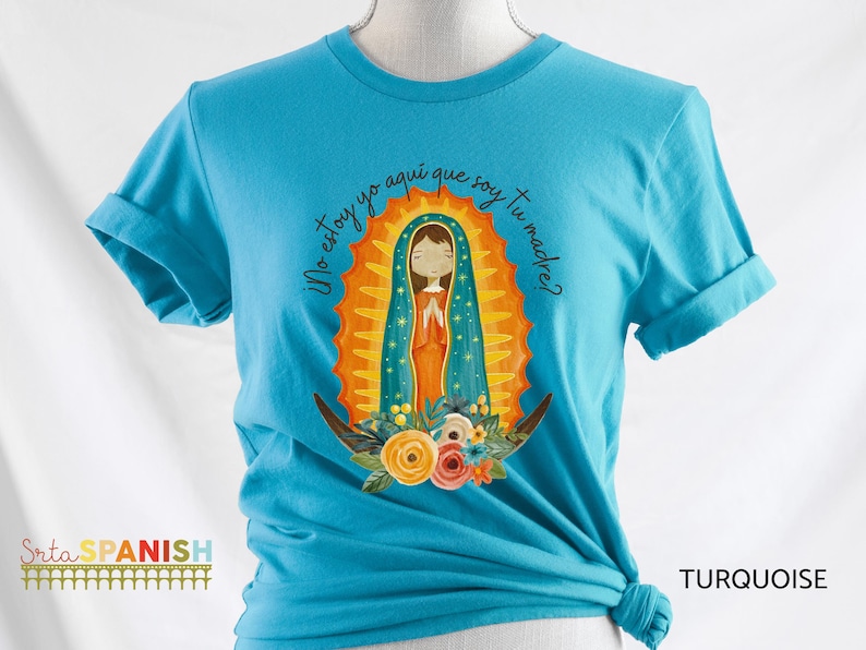 Puede incluir: Una camiseta azul turquesa con un gr&aacute;fico de la Virgen Mar&iacute;a en un halo dorado y azul. El texto "Yo estoy yo aqui que soy tu madre?" est&aacute; impreso en la camiseta.