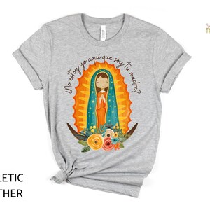 Puede incluir: Una camiseta gris jaspeada con un gr&aacute;fico de la Virgen Mar&iacute;a en una t&uacute;nica azul y dorada. El texto "&iexcl;No estoy yo aqu&iacute; que soy tu madre?" est&aacute; impreso en la camiseta.
