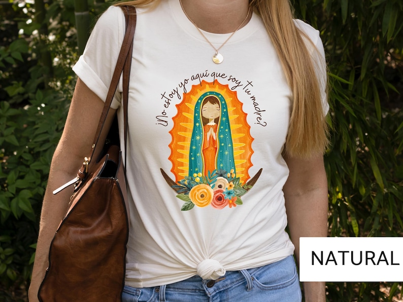 Puede incluir: Camiseta blanca con una ilustraci&oacute;n colorida de la Virgen Mar&iacute;a. El texto en la camiseta dice "&iexcl;No estoy yo aqu&iacute; que soy tu madre?"