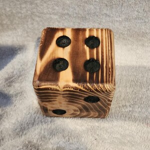 May include: A wooden dice with four black dots burned into the surface. The dice is made of light brown wood with dark brown wood grain.