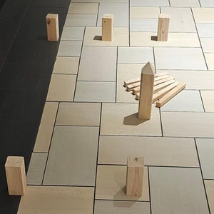 May include: An abstract composition featuring several small, rectangular wooden blocks and a central arrangement of wooden pieces. The blocks are arranged on a tiled surface with a mix of light and dark tones, creating a geometric pattern.