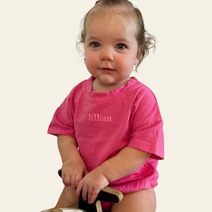 May include: A bright pink short-sleeved t-shirt featuring the name lillian embroidered in a matching pink thread on the center chest. The shirt has a crew neckline and a relaxed fit, worn by a toddler with light brown hair styled in a top knot.