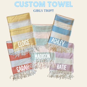 May include: Custom beach towels with names, featuring horizontal stripes in various colors: yellow, orange, blue, pink, and green. Each towel has a different name printed on it: Eloise, Sarah, Ashley, Madison, and Katie. The text "CUSTOM TOWEL" and "GIRLS TRIP!!!" are at the top.