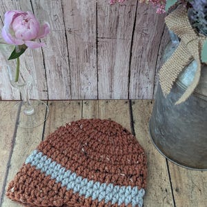 May include: A crochet beanie hat featuring a textured brown yarn with subtle white flecks and a single diagonal stripe of light gray yarn. The hat is constructed with a thick, visible stitch pattern and has a rounded, fitted shape.