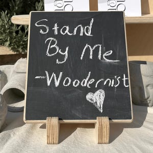 Könnte beinhalten: Eine kleine Tafel mit den Worten "Stand By Me - Woodernist" in weißer Kreide sowie einem Herzsymbol. Das Schild wird von einem Holzständer getragen. Der Hintergrund umfasst dekorative Gegenstände und bedruckte Karten.