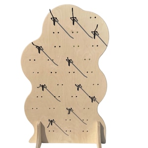 May include: A light-colored wooden display stand with a wavy top and two supporting legs. The stand has multiple small holes with black hooks, designed for hanging items. The wood has a natural grain and is set against a white background.
