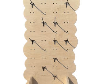 May include: A light-colored wooden display board with a scalloped edge, featuring a grid of pre-drilled holes and several attached metal hooks. The vertical panel is supported by two wooden feet at the base, allowing it to stand upright.