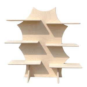 Puede incluir: Una estantería de pared de madera clara con un diseño único en forma de estrella. Cuenta con seis estantes rectangulares de diferentes tamaños, sostenidos por un panel trasero central y pequeñas patas. La madera tiene un aspecto natural sin terminar.