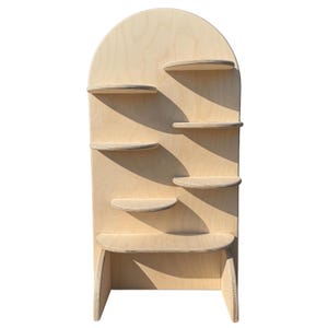 May include: A light-colored wooden wall-mounted display unit featuring an arched top panel and six staggered, semi-circular shelves of varying sizes. The natural wood grain is visible throughout the structure, which is supported by two side panels.