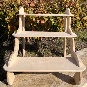May include: A three-tiered wooden shelf unit with rounded edges and a natural wood finish. The shelf is made of plywood and has a simple, modern design.