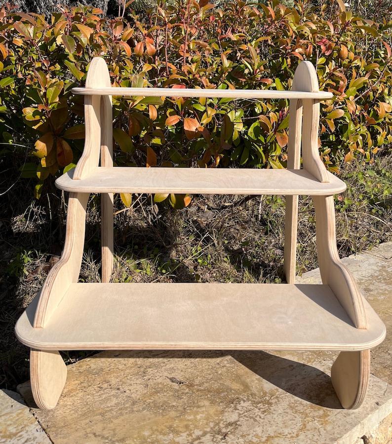 May include: A three-tiered wooden shelf unit with rounded edges and a natural wood finish. The shelf is made of plywood and has a simple, modern design.