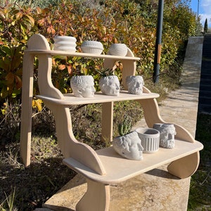 May include: A wooden shelf unit with three tiers, featuring a curved design. The shelves are decorated with white ceramic planters in the shape of human heads and other decorative planters.