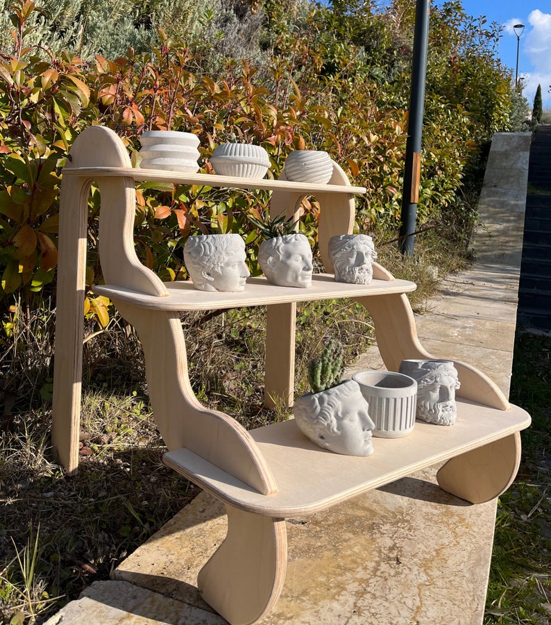 May include: A wooden shelf unit with three tiers, featuring a curved design. The shelves are decorated with white ceramic planters in the shape of human heads and other decorative planters.