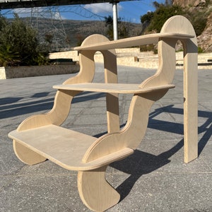 May include: A three-tiered wooden shelf unit with a unique, curved design. The shelves are made of light-coloured wood and have a smooth, natural finish.