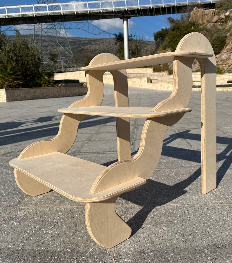 May include: A three-tiered wooden shelf unit with a unique, curved design. The shelves are made of light-coloured wood and have a smooth, natural finish.