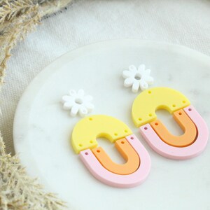 May include: A pair of colorful statement earrings featuring a yellow, orange, and pink layered arch shape with a white flower charm.