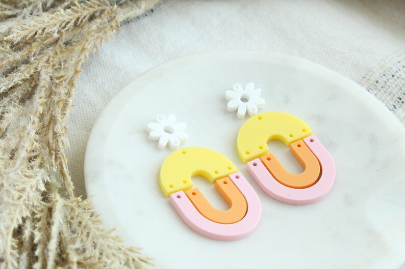 May include: A pair of colorful statement earrings featuring a yellow, orange, and pink layered arch shape with a white flower charm.