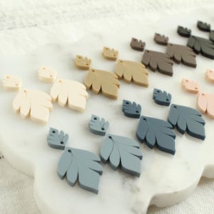 May include: A collection of leaf-shaped embellishments in various colors, including cream, tan, gray, and pink, arranged on a white, cloud-shaped surface. The leaves vary in size and are likely for crafting or decorative purposes.