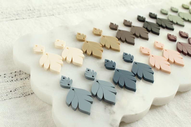 May include: A collection of leaf-shaped embellishments in various colors, including cream, tan, gray, and pink, arranged on a white, cloud-shaped surface. The leaves vary in size and are likely for crafting or decorative purposes.