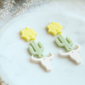 May include: A pair of earrings featuring a yellow sun, a green cactus, and a white bull's head charm. The earrings are made of polymer clay and are perfect for adding a touch of western style to any outfit.