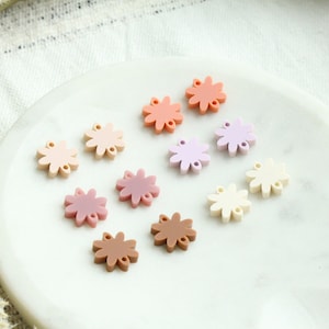 May include: Assortment of ten flower-shaped resin charms in various pastel colors, including pink, peach, lavender, and cream. The charms are arranged on a white surface.