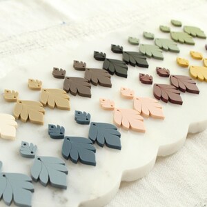 May include: An assortment of leaf-shaped decorative elements in various colors, including cream, tan, gray, blue, pink, brown, yellow, and green. The leaves are arranged on a white, scalloped-edge surface, creating a visually appealing display.