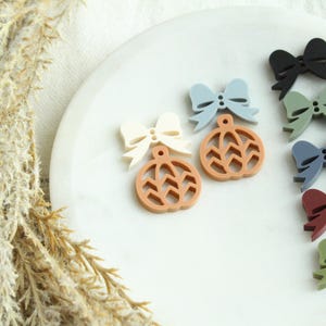 May include: A collection of colorful bow-shaped ornaments in various shades, including black, green, blue, and red, paired with pumpkin-shaped ornaments in cream and brown. The ornaments are arranged on a white plate.
