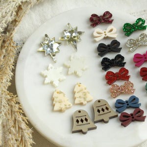 May include: A collection of Christmas-themed embellishments, including star and snowflake shapes, miniature Christmas trees, and bell shapes. The assortment also features various bow shapes in colors like red, green, and black, with glitter accents.