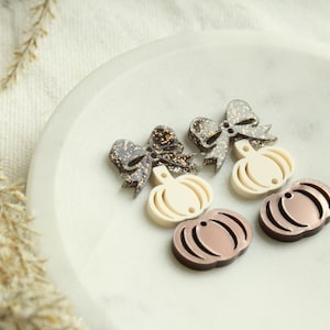 May include: A collection of fall-themed acrylic charms. The charms include two bow shapes with glitter accents, two cream-colored pumpkins, and two brown pumpkins. The charms are arranged on a white, circular surface.