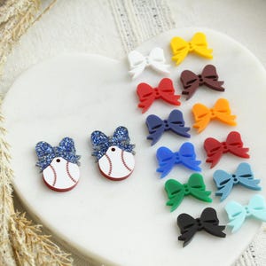 May include: A collection of colorful bow-shaped accessories in various colors, including white, yellow, red, brown, orange, blue, green, and black. Two baseball-themed accessories with glittery blue bows are also present. The items are arranged on a white, heart-shaped surface.